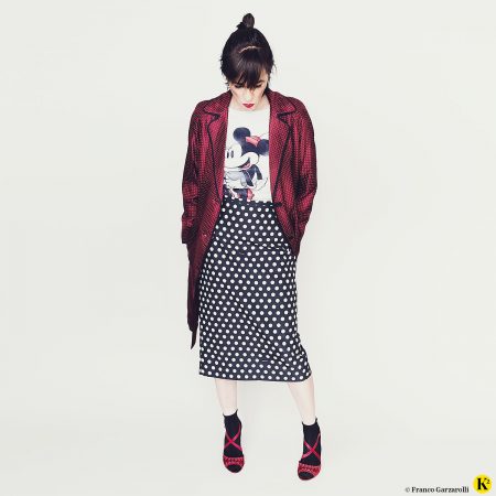 Karin trägt einen schwarzen Bleistiftrock mit weißen Punkten plus Minnie-Maus-Sweater und Seidenmantel in Bordeaux.