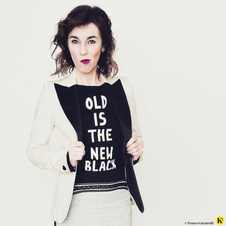 Karin trägt ein schwarzes T-Shirt mit der Aufschrift "Old is the new black" und schaut lustig in die Kamera. Weiters trägt sie einen cremefarbigen Rock und ein cremefarbiges Blouson.