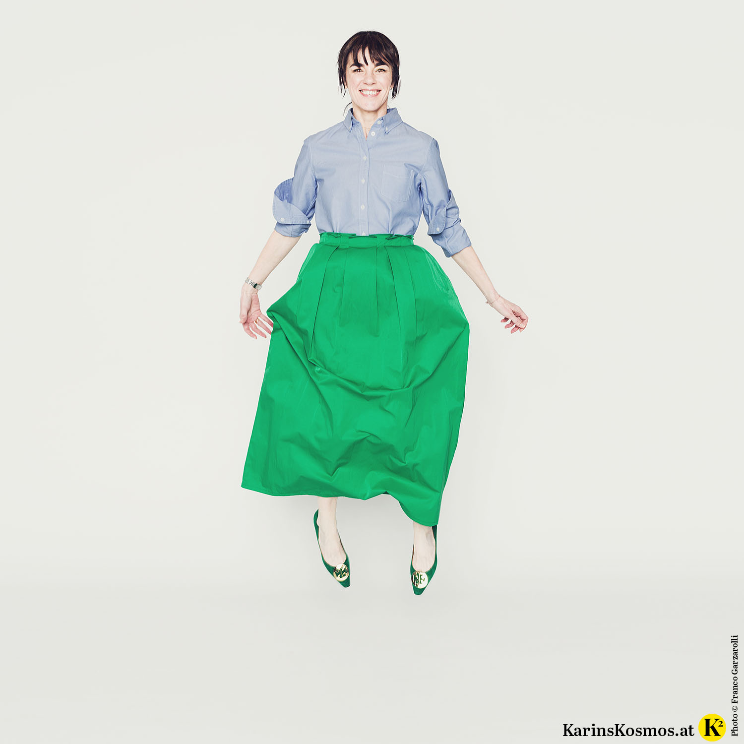 Frau springt in hellblauer Button Down-Bluse mit grünem, langem Rock und grünen Ballerinas.