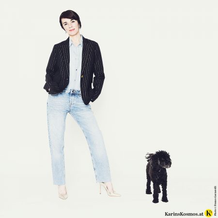 Frau trägt Jeanshemd und Jeans zu schwarzem Blazer und goldenen Schuhen. Daneben ein schwarzer Pudel.