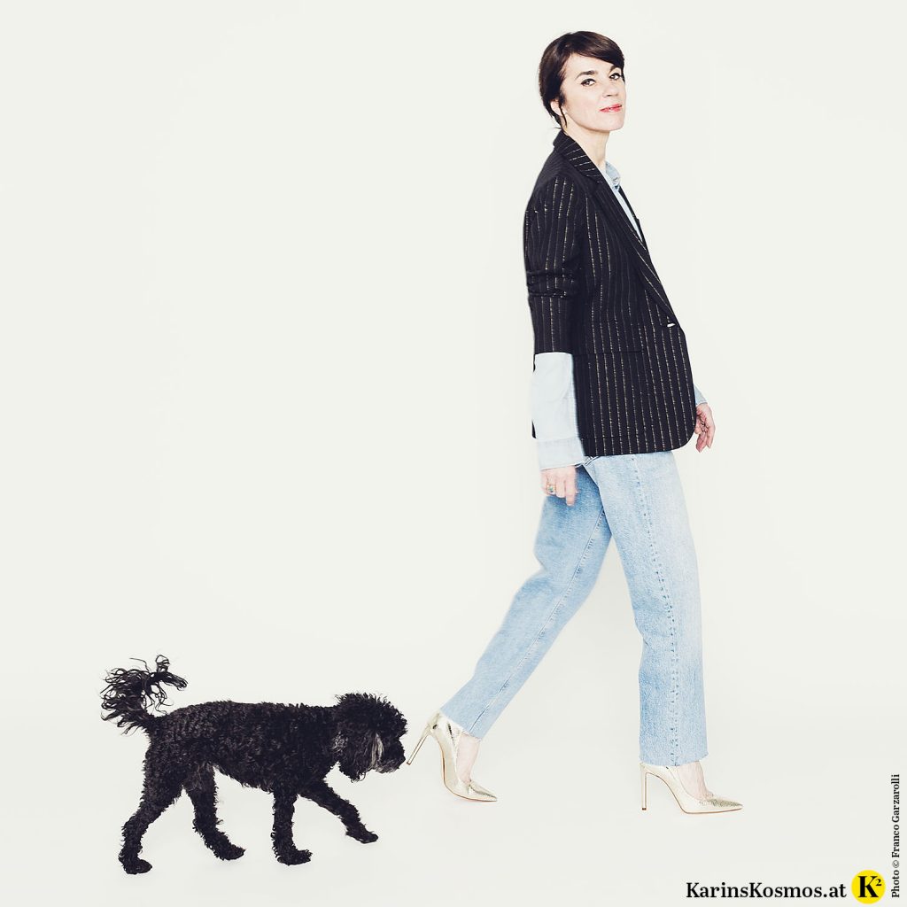 Frau geht in Jeans-Outfit mit dunklem Blazer und hinter ihr ein schwarzer Toypudel.