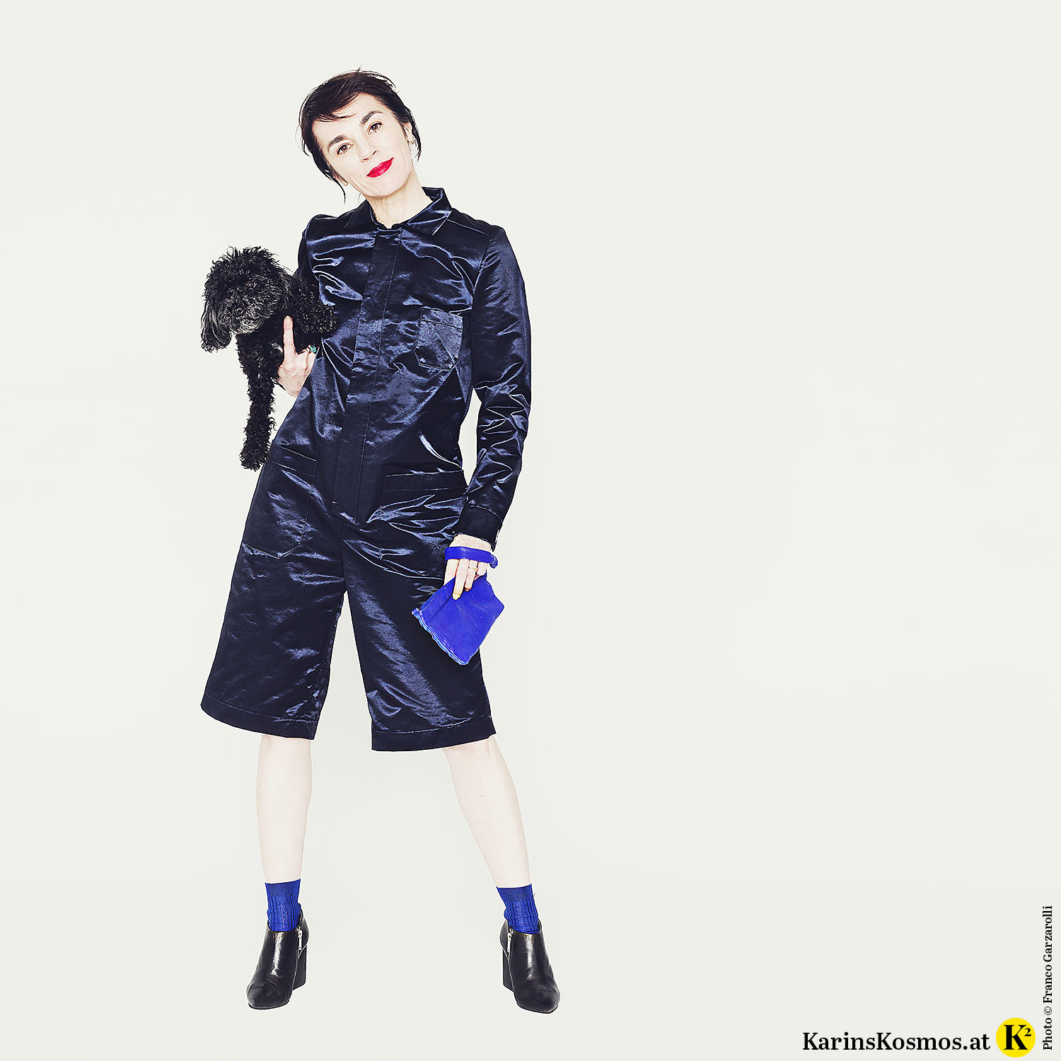 Frau mit Toypudel Trudy am Arm in mitternachtsblauem Overall mit Clutch und Ankle-Boots.