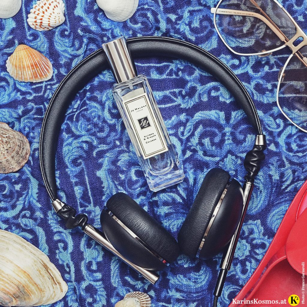Stilllife mit Parfumflakon von Jo Malone, Nutmeg & Ginger, Kopfhörer, Muscheln, Sonnenbrillen auf blauem Tuch.