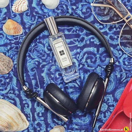 Stilllife mit Parfumflakon von Jo Malone, Nutmeg & Ginger, Kopfhörer, Muscheln, Sonnenbrillen auf blauem Tuch.