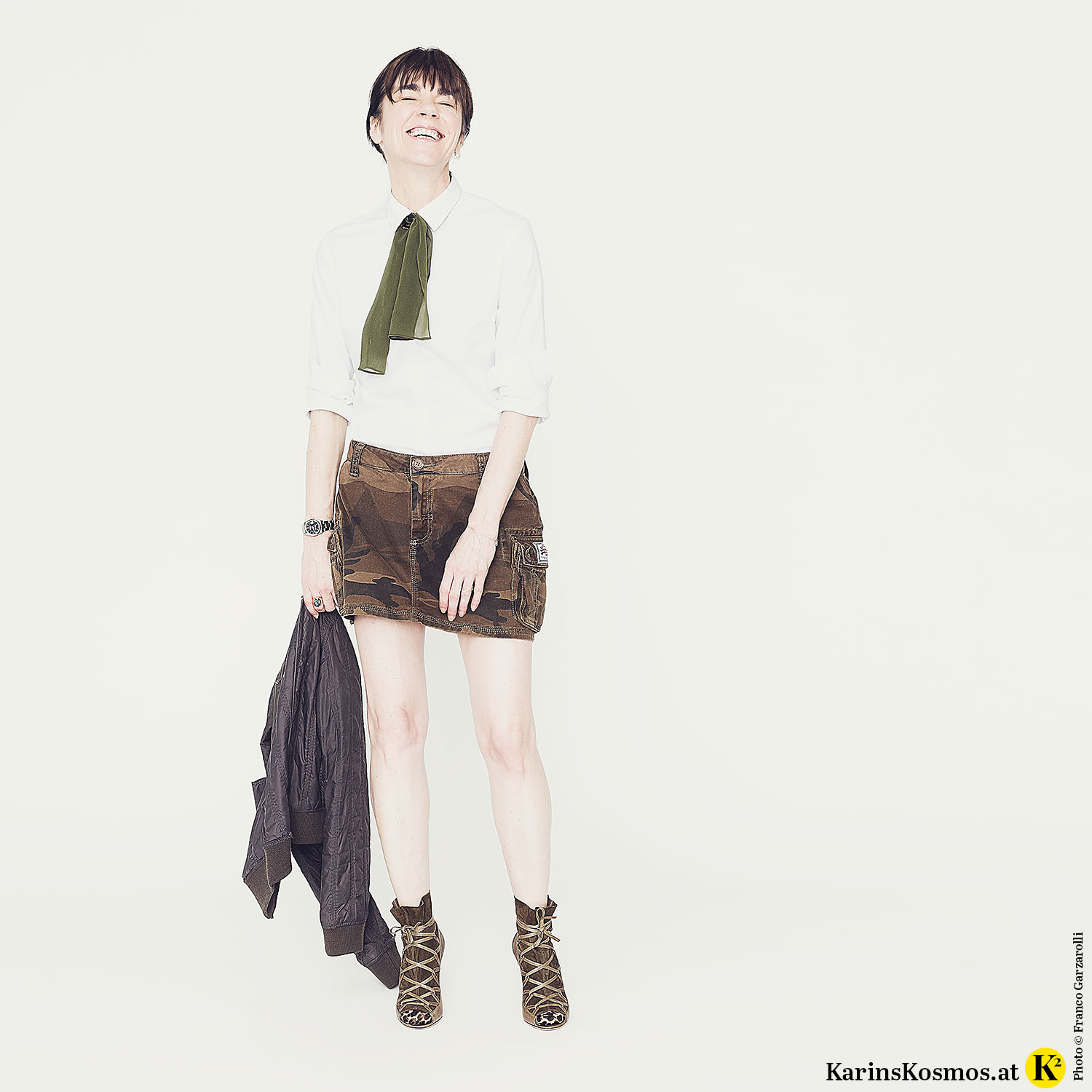 Frau in Minirock mit weißer Bluse, Seidenschal, Bomberjacke und High-Heels.