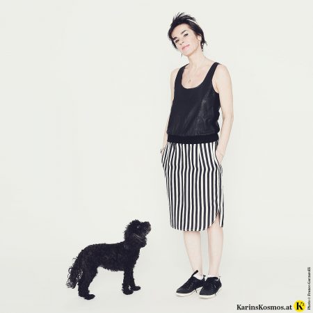 Frau in schwarzem Top, Seidenrock mit schwarz/weißen Streifen und schwarzen Sneakers. Vor ihr steht ein schwarzer Toypudel.