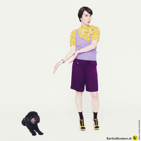 Frau mit gelb-gemusterter Bluse, lila Pullunder, violetter Short und gelben Sandalen. Daneben Toypudel der gähnt und Farben nicht so sieht.