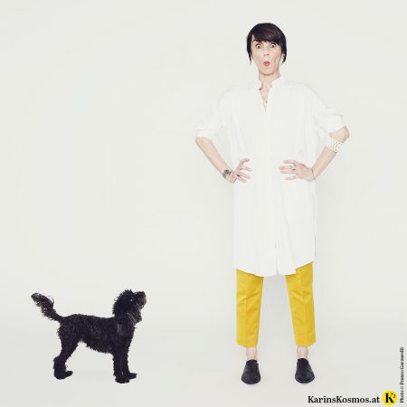 Frau in weißem Hemdkleid mit gelber Seidenhose und schwarzen Schuhen. Daneben steht ein schwarzer Toypudel.
