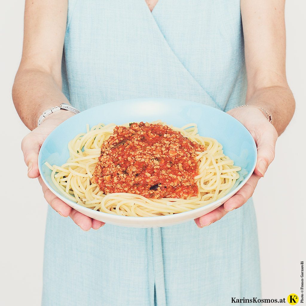 Frau in einem Türkisen Wickelkleid hält einen Teller mit Spaghetti Bolognese.