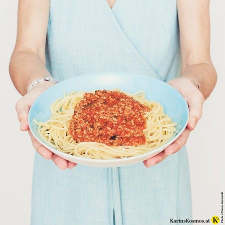 Frau in einem Türkisen Wickelkleid hält einen Teller mit Spaghetti Bolognese.
