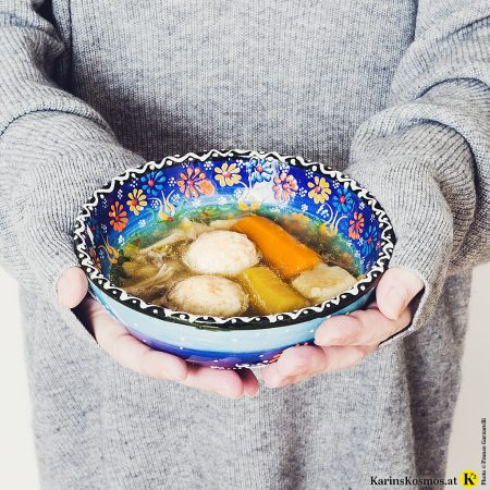In diesem Bild hält eine Frau, die einen grauen Pulli anhat, eine bunte Schüssel mit Hühnersuppe darin.
