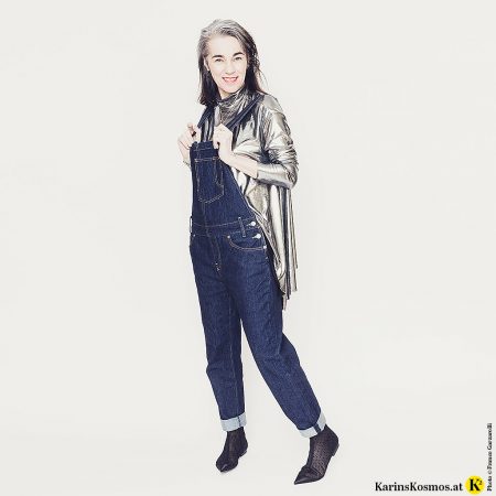 Frau in einer Jeanslatzhose, dazu trägt sie ein silberfarbenes Top und Stoff-Stiefeletten.